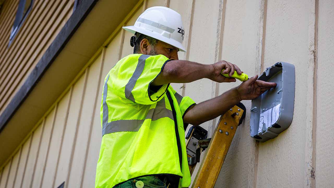 Ziply Fiber technician stands on ladder to install an Optical Network Terminal (ONT) on the side of a home