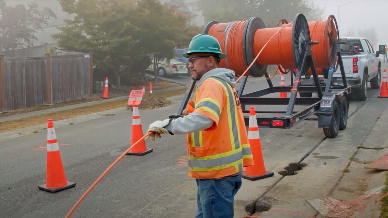 Technician in hard hat pulls cable from a truck on a road marked by traffic cones