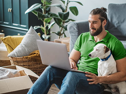 Person sitting on floor in front of an open moving box, with dog and computer on lap